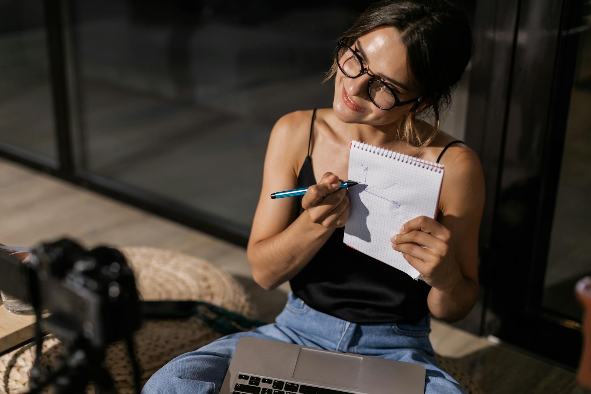 Smiling woman vlogging indoors with a camera, laptop, and notepad.